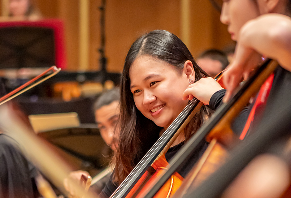 A young Asian woman smiles as she plays the cello with the RCM Symphony Orchestra.